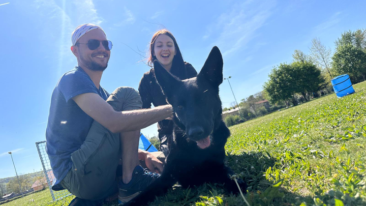 Schwarzer Schäferhund liegt auf der Wiese zwischen zwei lachenden Menschen an einem sonnigen Tag.