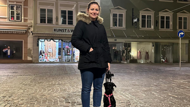 Frau mit kleinem schwarzen Hund an der Leine auf einem beleuchteten Stadtplatz am Abend.